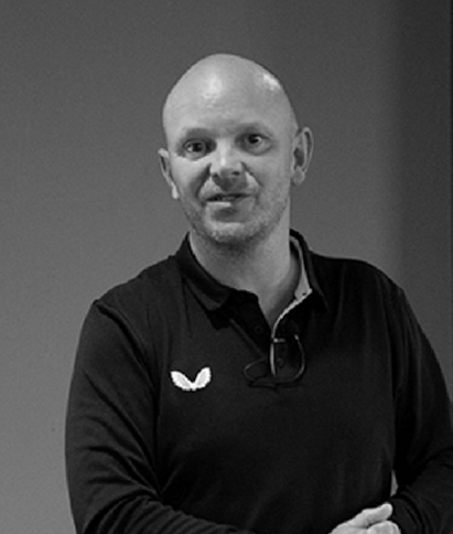 A bald man wearing a black shirt and glasses is standing with his arms crossed.