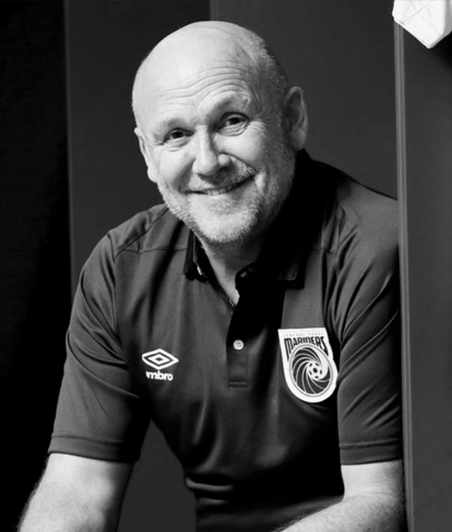 A man with a beard is leaning against a wall and smiling in a black and white photo.
