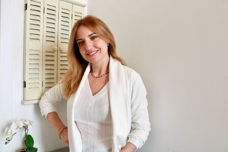 Una mujer vestida de blanco sonríe, parada cerca de una pared con una contraventana y una flor.