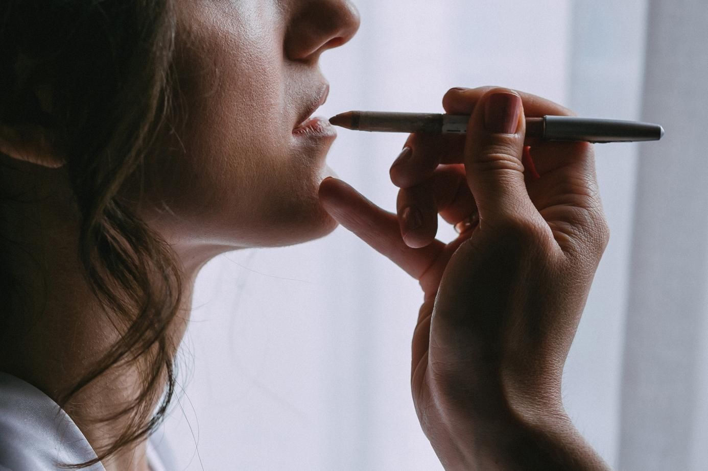 Mujer aplicando delineador de labios con un lápiz cerca de una ventana.