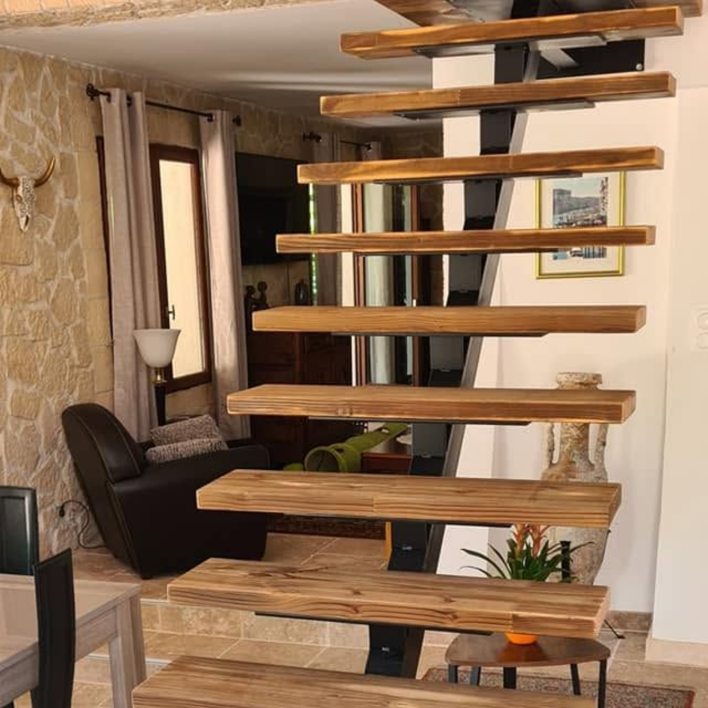 Salon moderne avec un escalier en bois suspendu, un mur en pierre et un fauteuil en cuir foncé.
