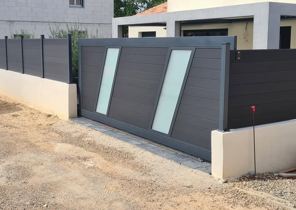 Portail d'entrée gris moderne avec des panneaux en verre dépoli, jouxtant un mur beige sur une allée de gravier.
