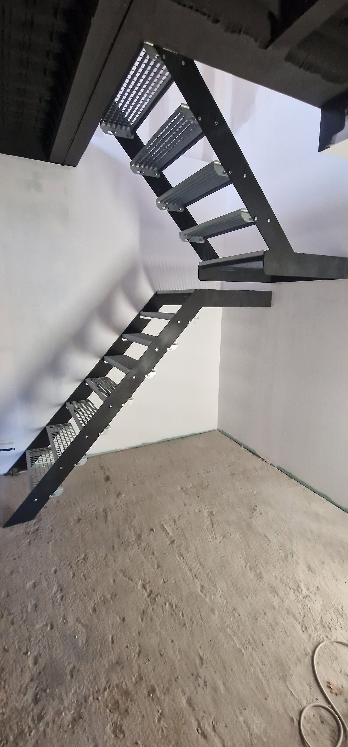 Escalier intérieur avec marches et rampes en métal noir dans une pièce blanche inachevée.