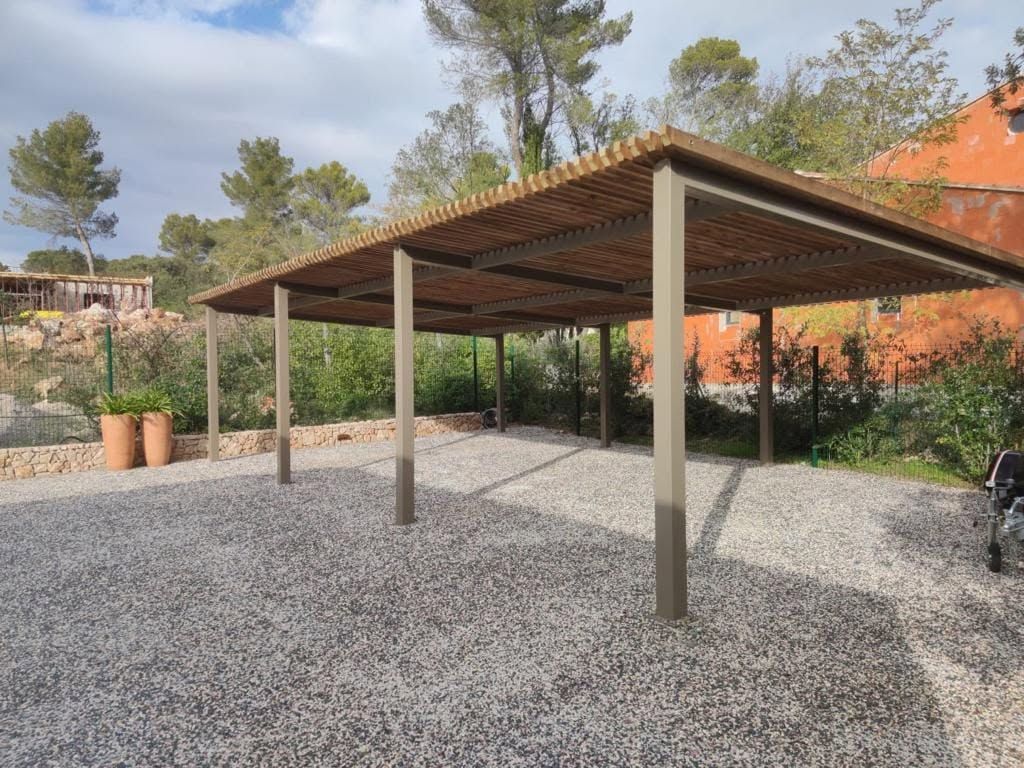 Tonnelle en bois et métal sur une terrasse en gravier, entourée d''une haie et de plantes en pot.