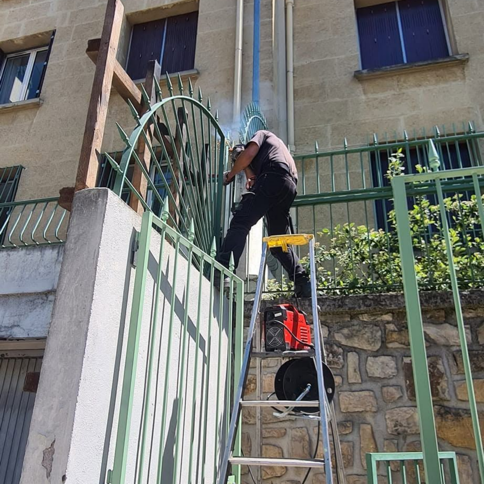 Un ouvrier, juché sur une échelle, répare un portail métallique vert devant un bâtiment en pierre.