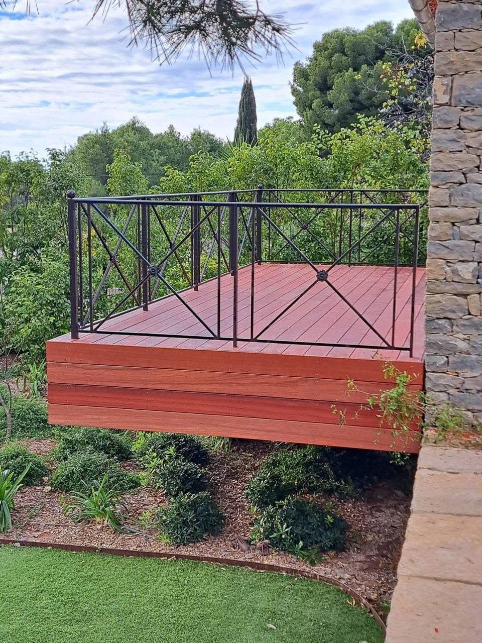 Balcon en bois rouge avec rambarde noire donnant sur un jardin verdoyant et des arbres.