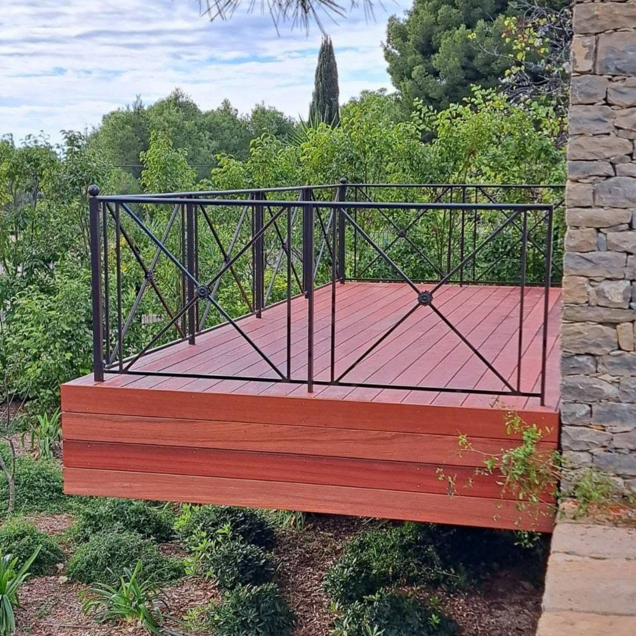 Balcon en bois rouge avec une rambarde noire, adossé à un mur de pierre, donnant sur une dense végétation.