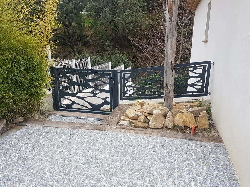 Allée pavée de dalles grises, portail en métal noir, aménagement paysager en pierre et mur d'enceinte blanc.