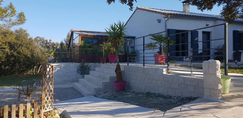 Maison blanche avec une terrasse en pierre, des balustrades noires et des plantes en pot roses.
