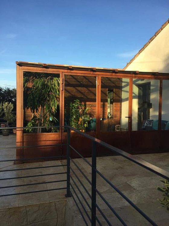 Patio de maison moderne avec structure en bois et en verre, rampe d'accès et plantes en pot baigné de soleil.