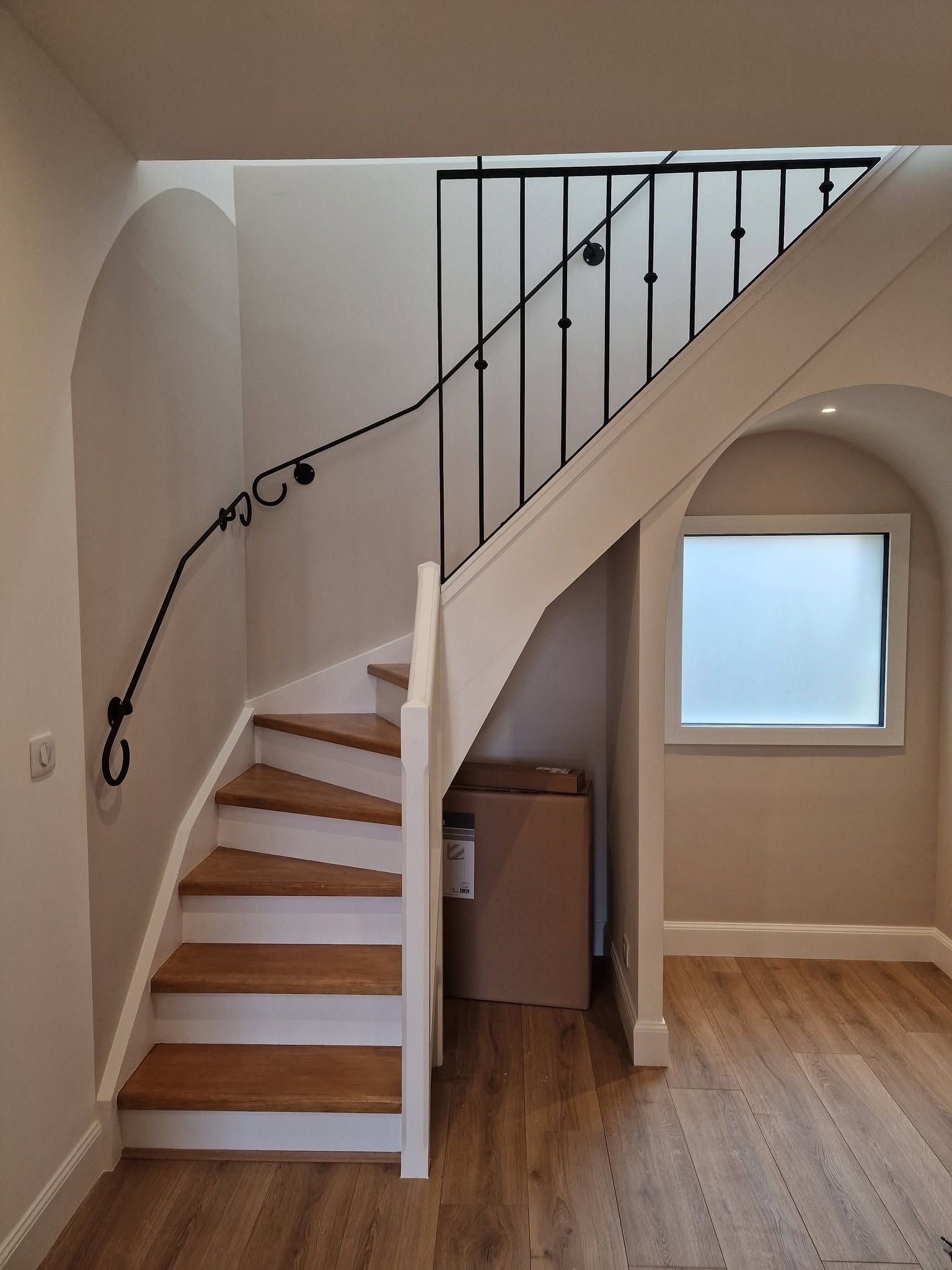 Escalier intérieur avec des marches en bois, une rampe en acier noir et petite fenêtre dans un couloir beige.