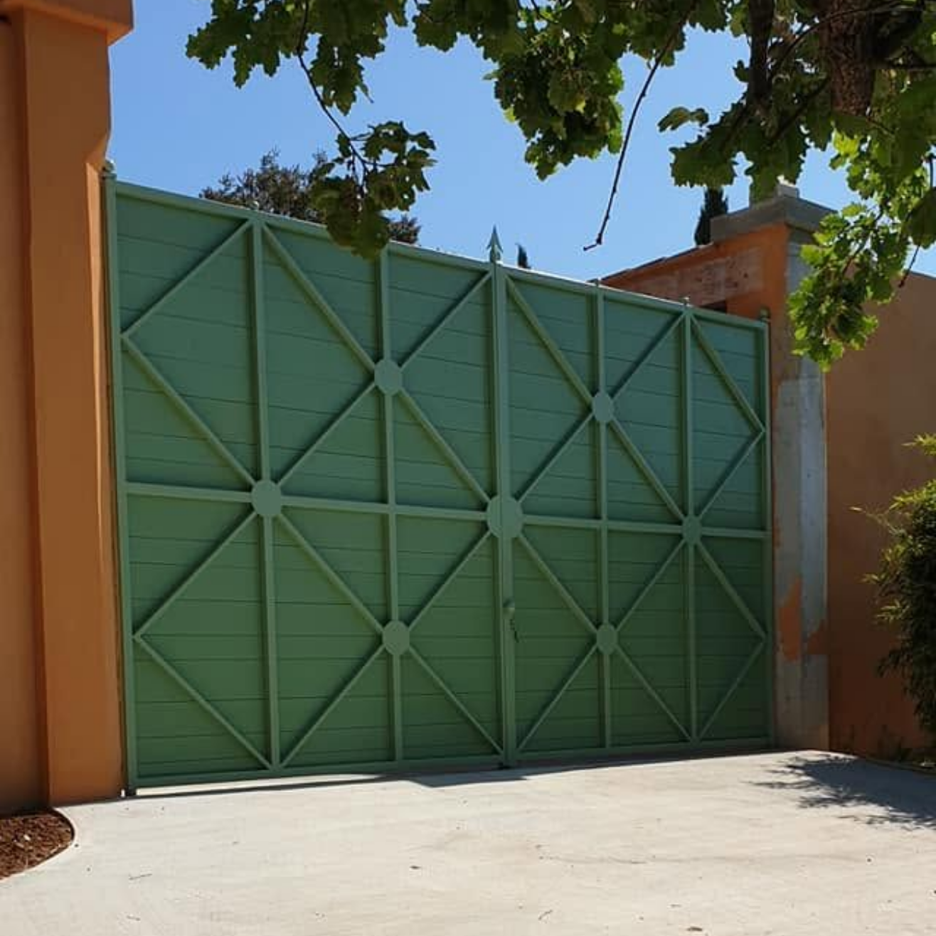 Un grand portail en métal vert est installé entre des murs beiges dans une allée ensoleillée.