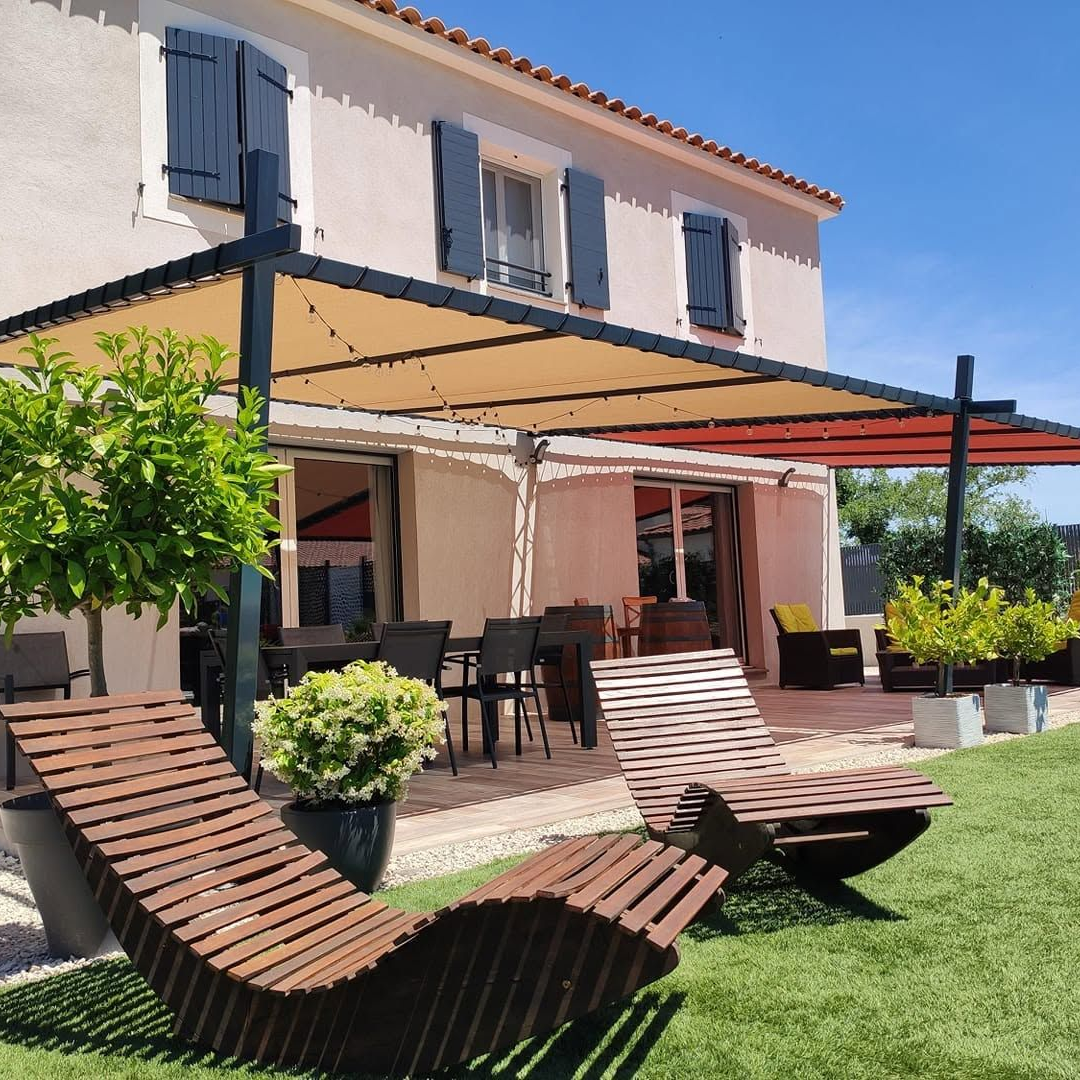 Maison à deux étages avec volets bleus et une terrasse ombragée sous une pergola avec des chaises longues rayées.