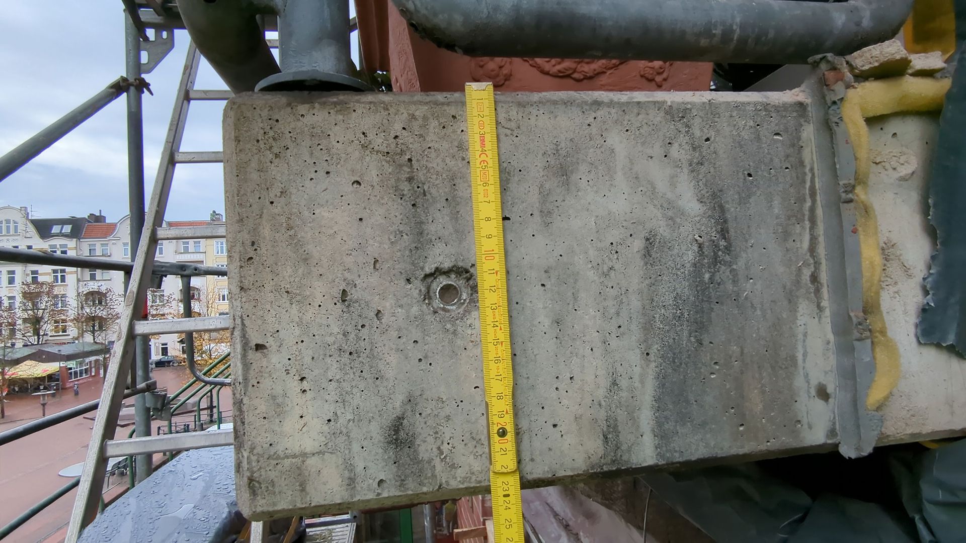 Betonblock auf einer Baustelle mit gelbem Maßband auf einem Gerüst, im Hintergrund Gebäude zu sehen.