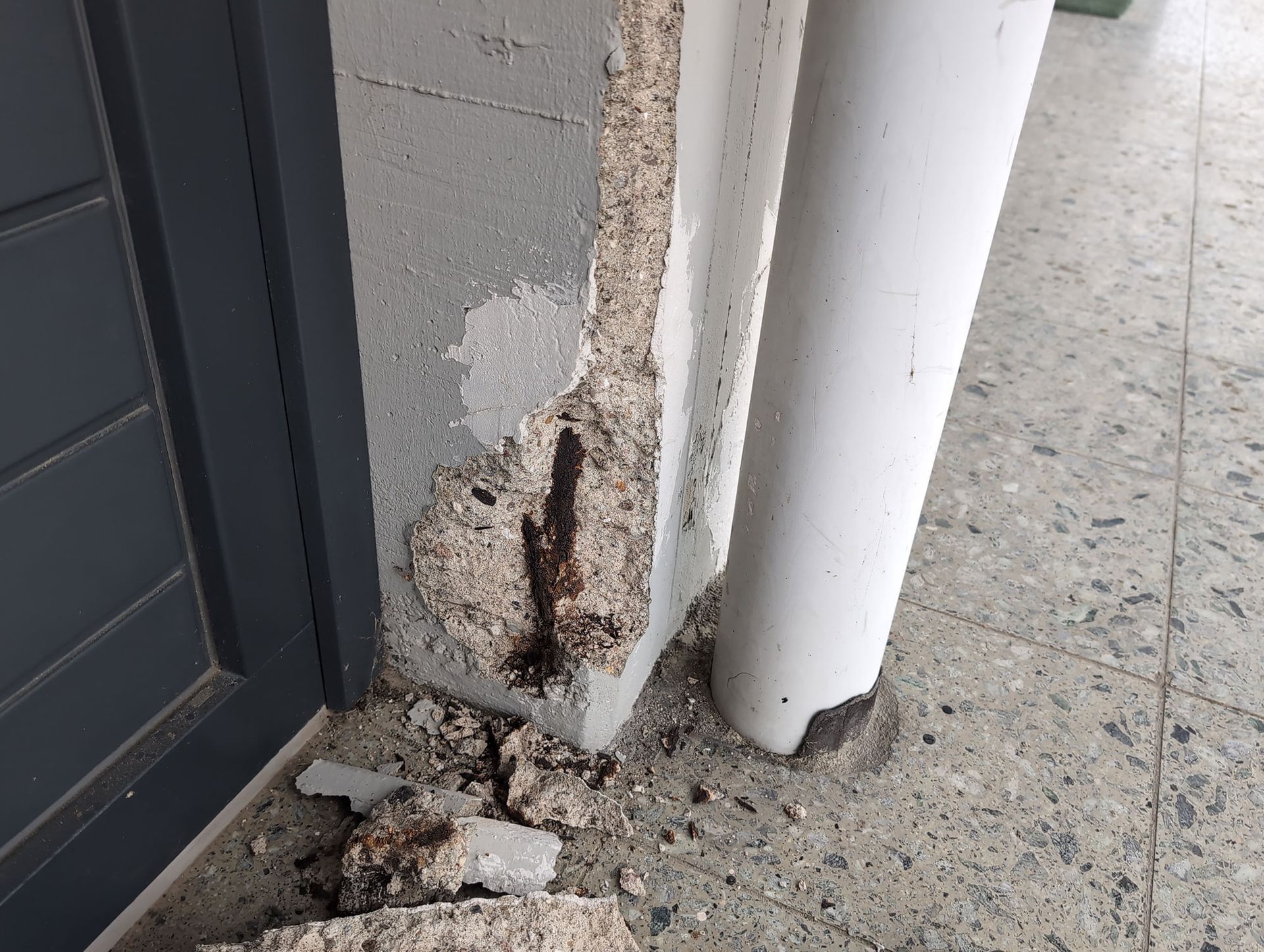 Beschädigter Mauersockel mit bröckelndem Beton und freiliegendem Rost neben einem Türrahmen und einer weißen Säule