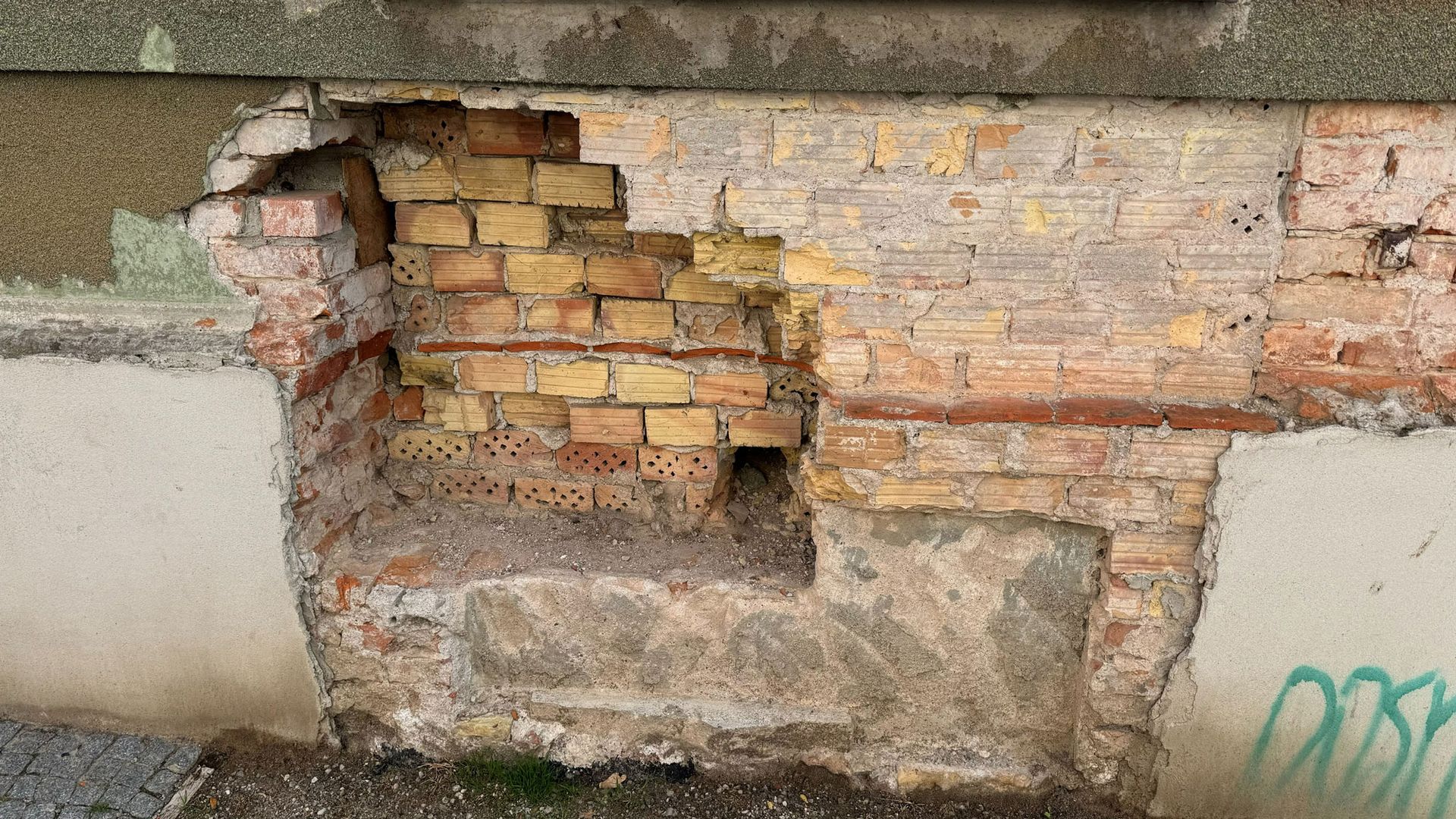 Beschädigte Ziegelmauer mit bröckelndem Putz und freiliegenden Ziegeln in der Nähe eines gestrichenen Betonsockels.