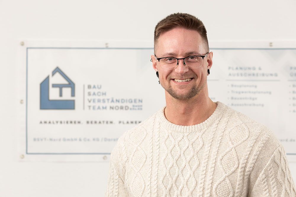 Ein lächelnder Mann in einem weißen Pullover steht vor einem Team-Branding-Schild in einer Büroumgebung.