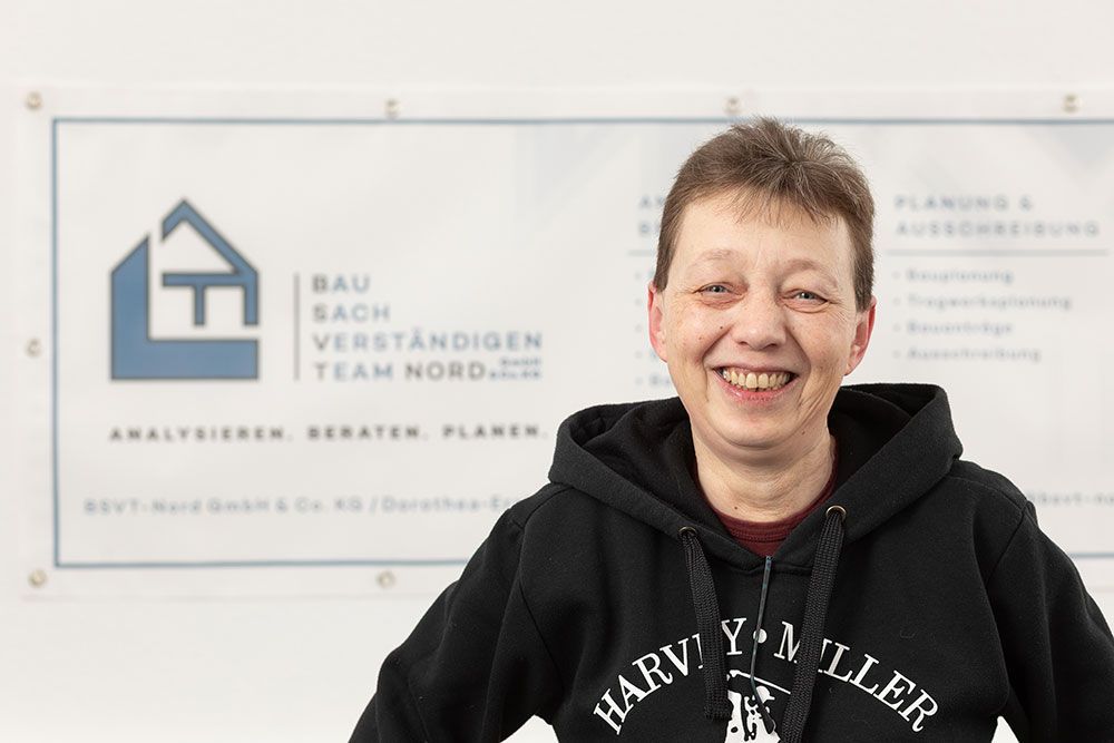 Eine lächelnde Person in einem schwarzen Kapuzenpullover steht vor einem Schild des BAU-Vastgoed-Teams.