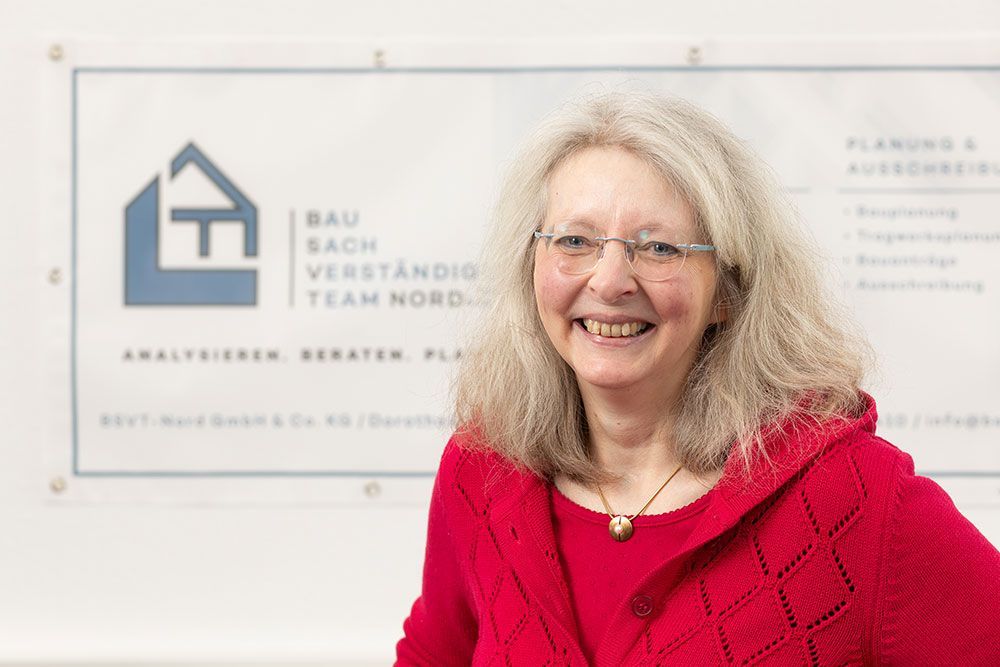 Eine lächelnde Frau in einem roten Pullover steht vor einem Wandschild mit der Aufschrift „Teamraum der Bauhaus-Universität“.