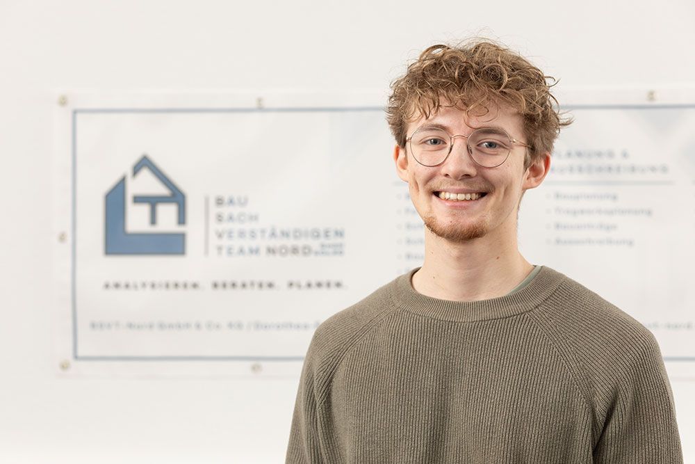 Eine lächelnde Person mit Brille steht vor einem verschwommenen Büroschild mit einem Hauslogo.