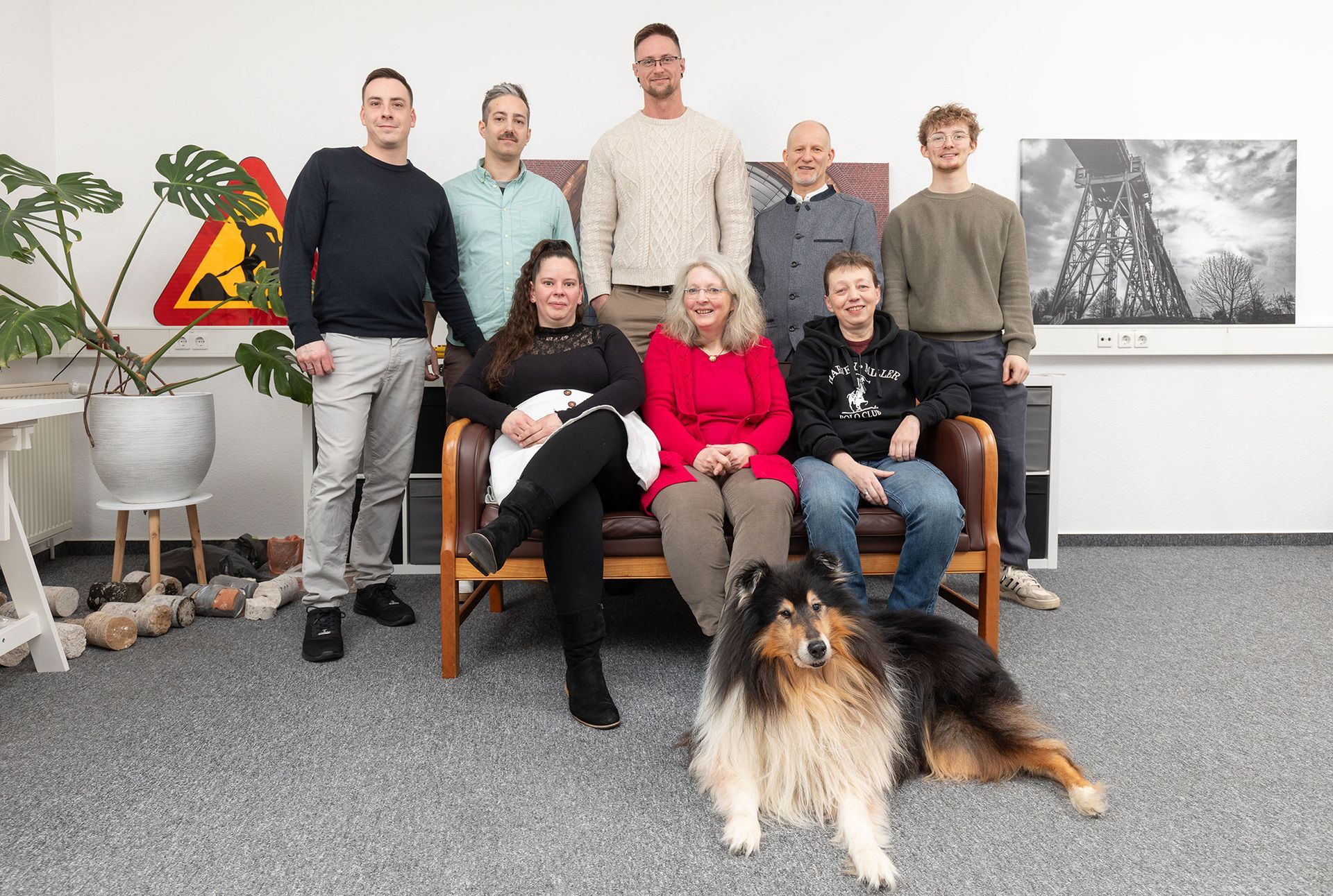 Eine Gruppe von sechs Personen posiert in einem Innenraum mit einem Collie-Hund vor sich.