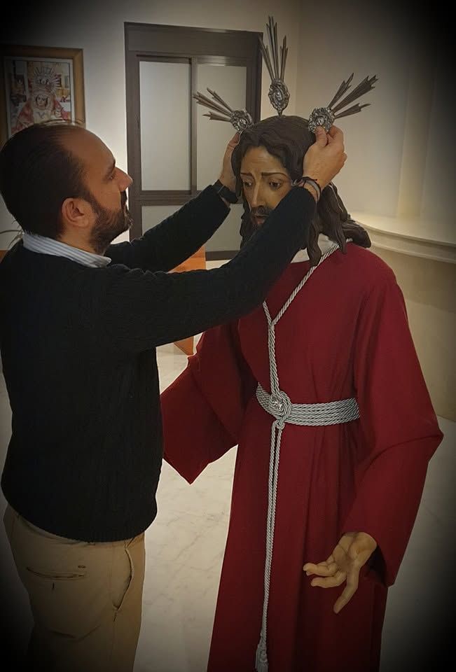 Hombre ajustando una estatua coronada o un maniquí con túnica roja en el interior de un edificio.