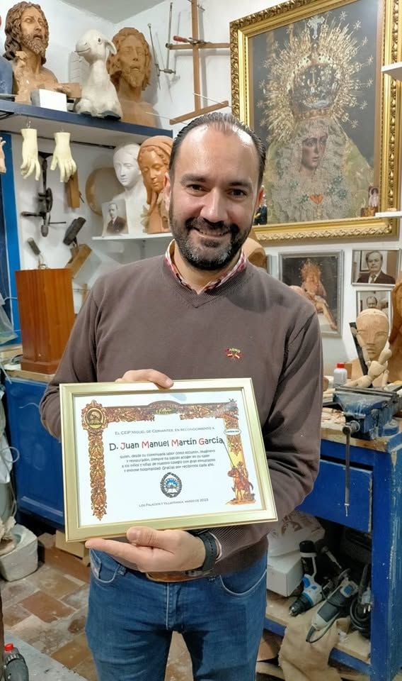 Un hombre sonríe en un estudio de arte, sosteniendo un certificado enmarcado, rodeado de esculturas