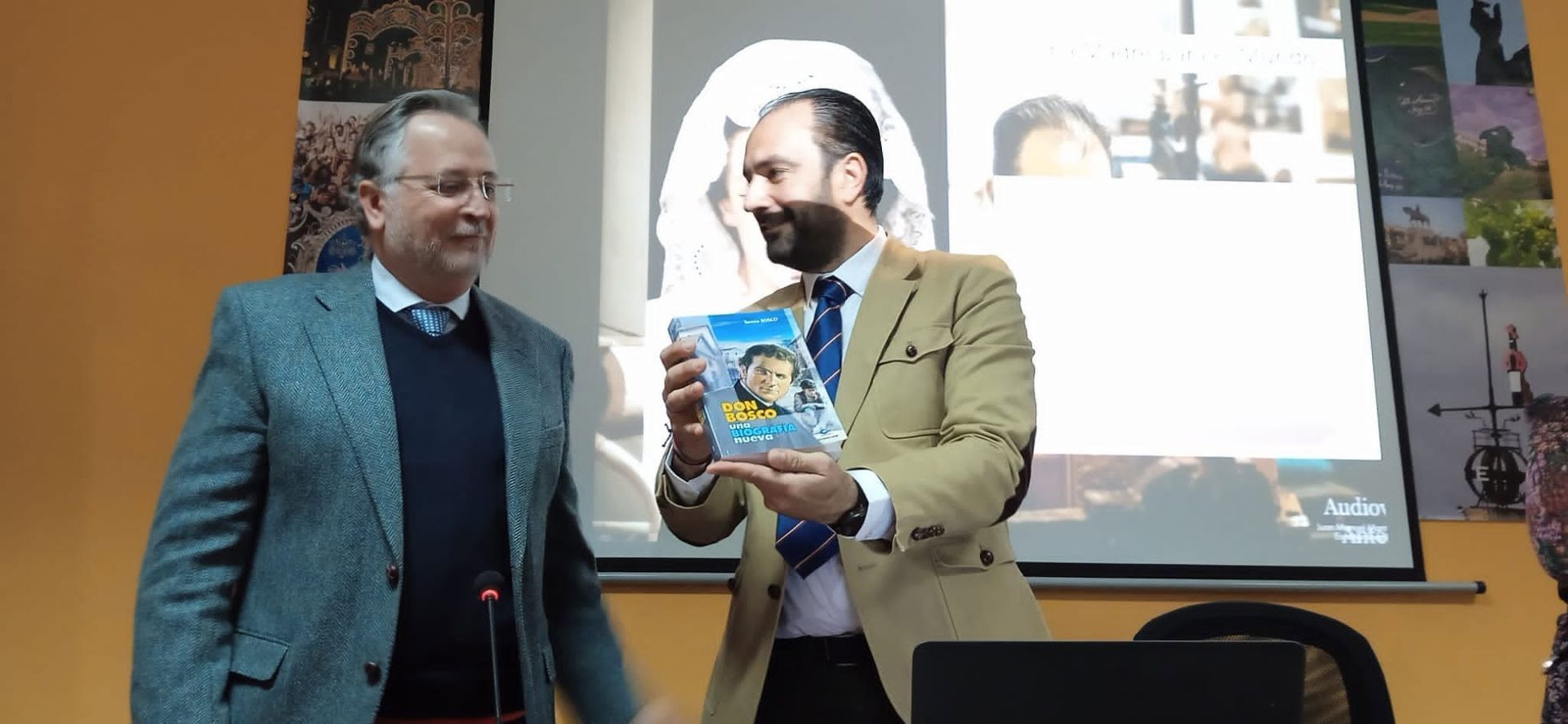 Dos hombres en el escenario, uno sosteniendo un libro colorido, con una diapositiva de presentación