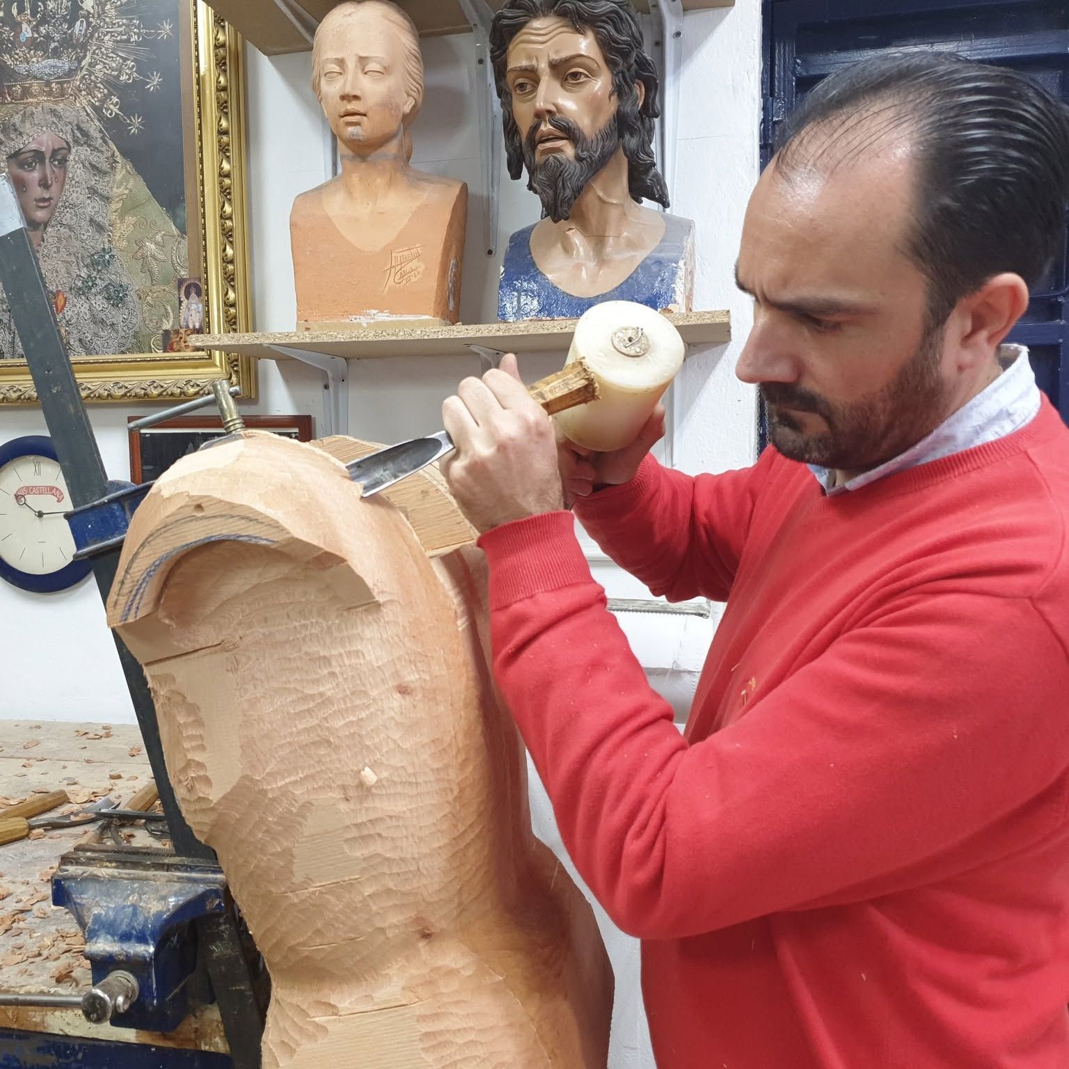 Un hombre con un suéter rojo talla una escultura de madera en un taller, con dos bustos expuestos detrás de él.