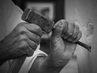Manos sujetando un martillo y un cincel contra una superficie de piedra, en blanco y negro.