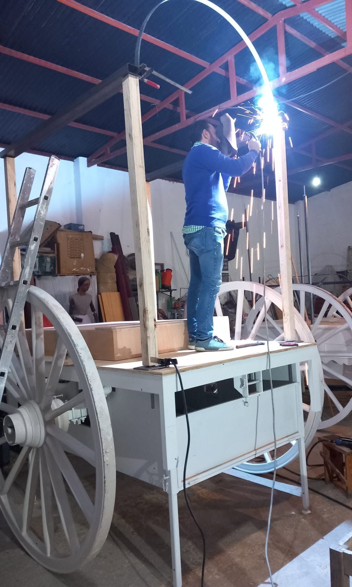 Un trabajador suelda una gran estructura metálica con ruedas en un taller, con brillantes chispas en el aire.