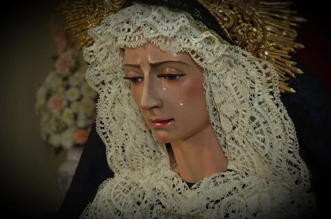 Estatua religiosa serena con velo de encaje, corona floral y una lágrima en la mejilla, en penumbra.