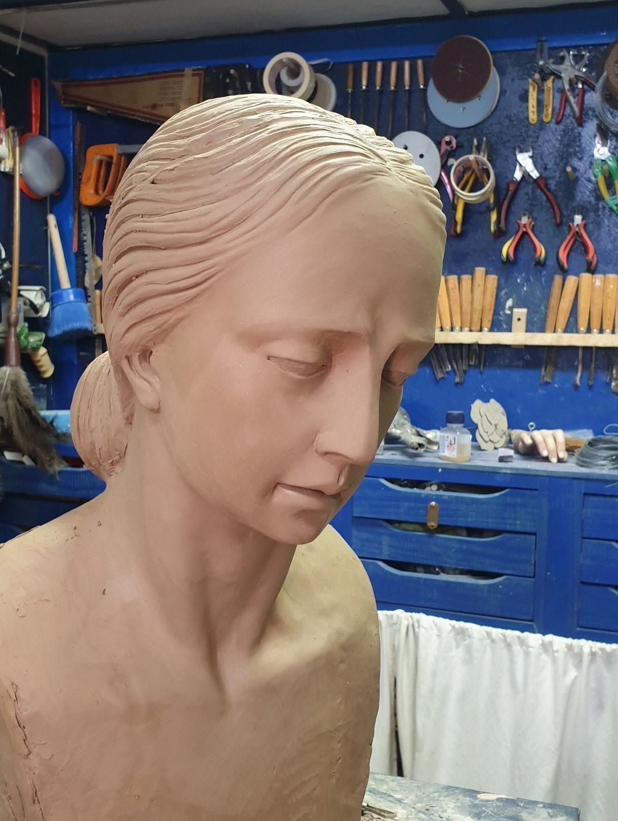 Busto de arcilla de una mujer de perfil sobre un banco de trabajo, con herramientas y cajones azules al fondo.