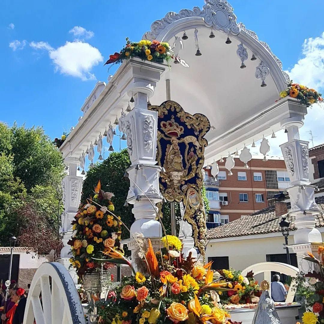 Carroza religiosa decorada bajo un arco blanco, cubierta de coloridas flores contra un cielo azul.
