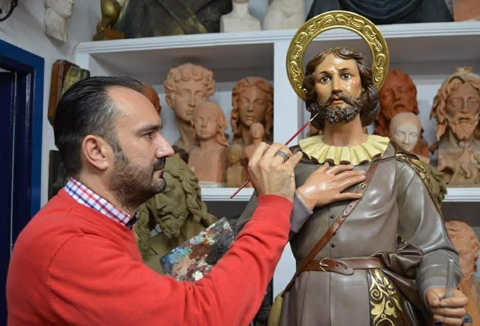 Hombre con suéter rojo pintando una estatua de un santo con aureola en un taller.