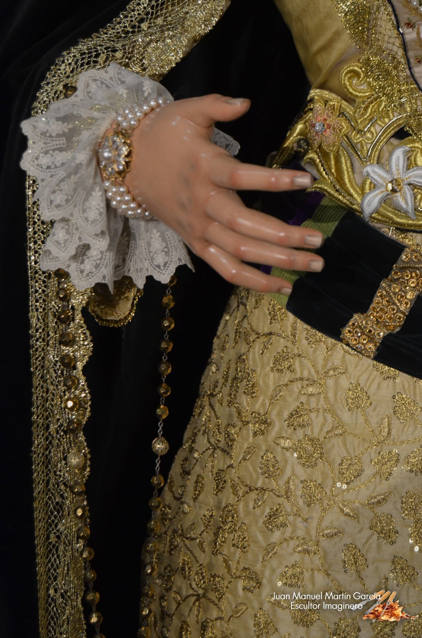 Primer plano de una mano con brazalete enjoyado y puño de encaje sobre tela negra bordada en oro.