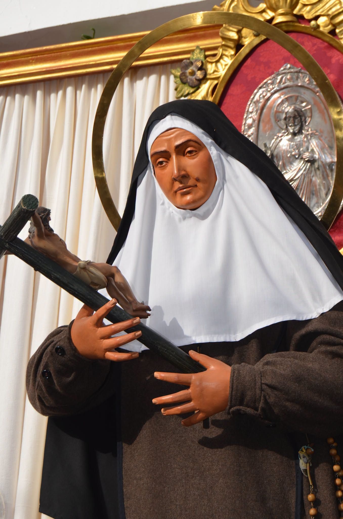 Estatua de una monja sosteniendo un crucifijo frente a un fondo decorativo dorado y rojo.