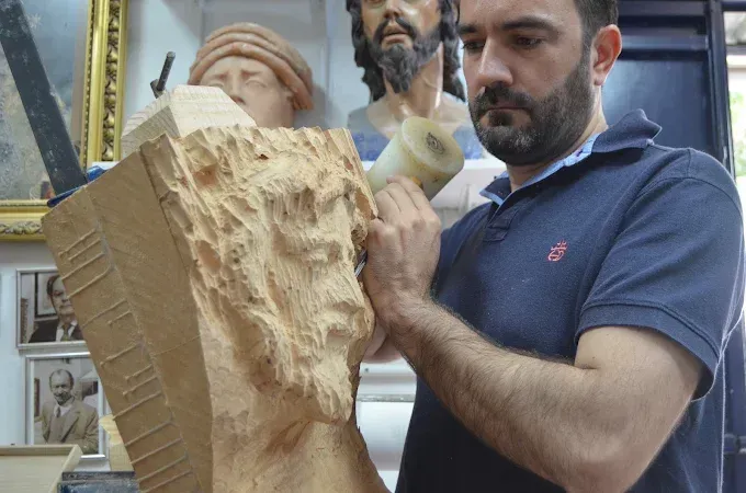 Un artista talla un busto de madera en un taller, con esculturas terminadas al fondo.
