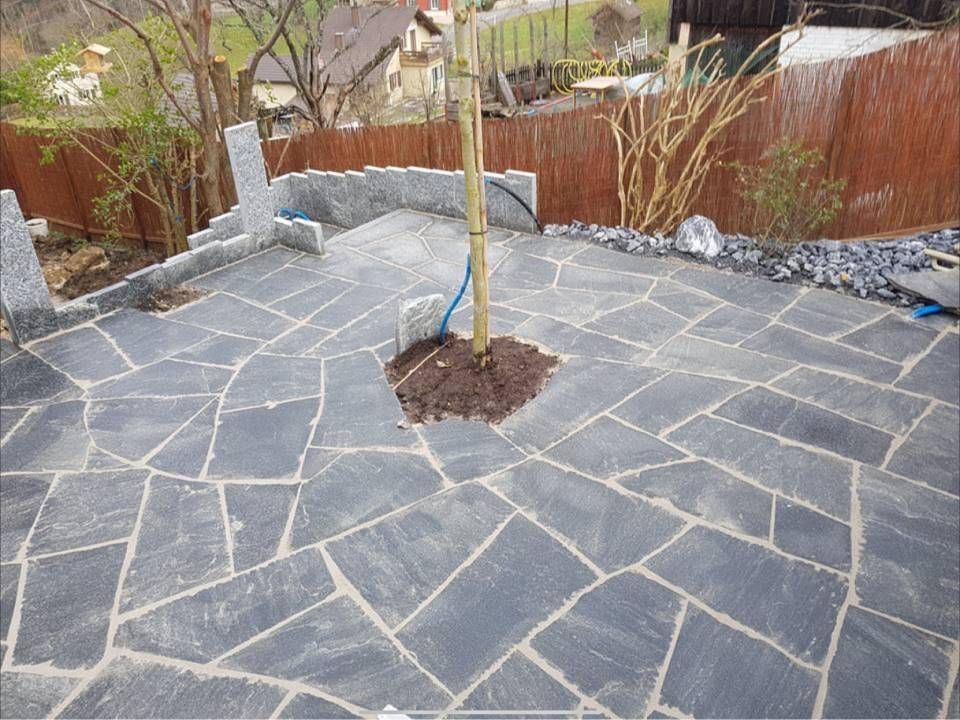 Neu gepflasterte Steinterrasse mit einem jungen Baum in der Mitte und einem umzäunten Garten im Hintergrund