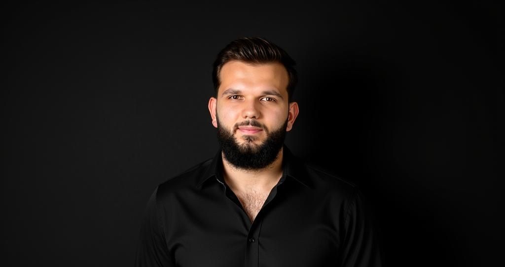 Ein Mann in schwarzem Hemd steht vor dunklem Hintergrund, den Blick geradeaus gerichtet, mit neutralem Gesichtsausdruck.