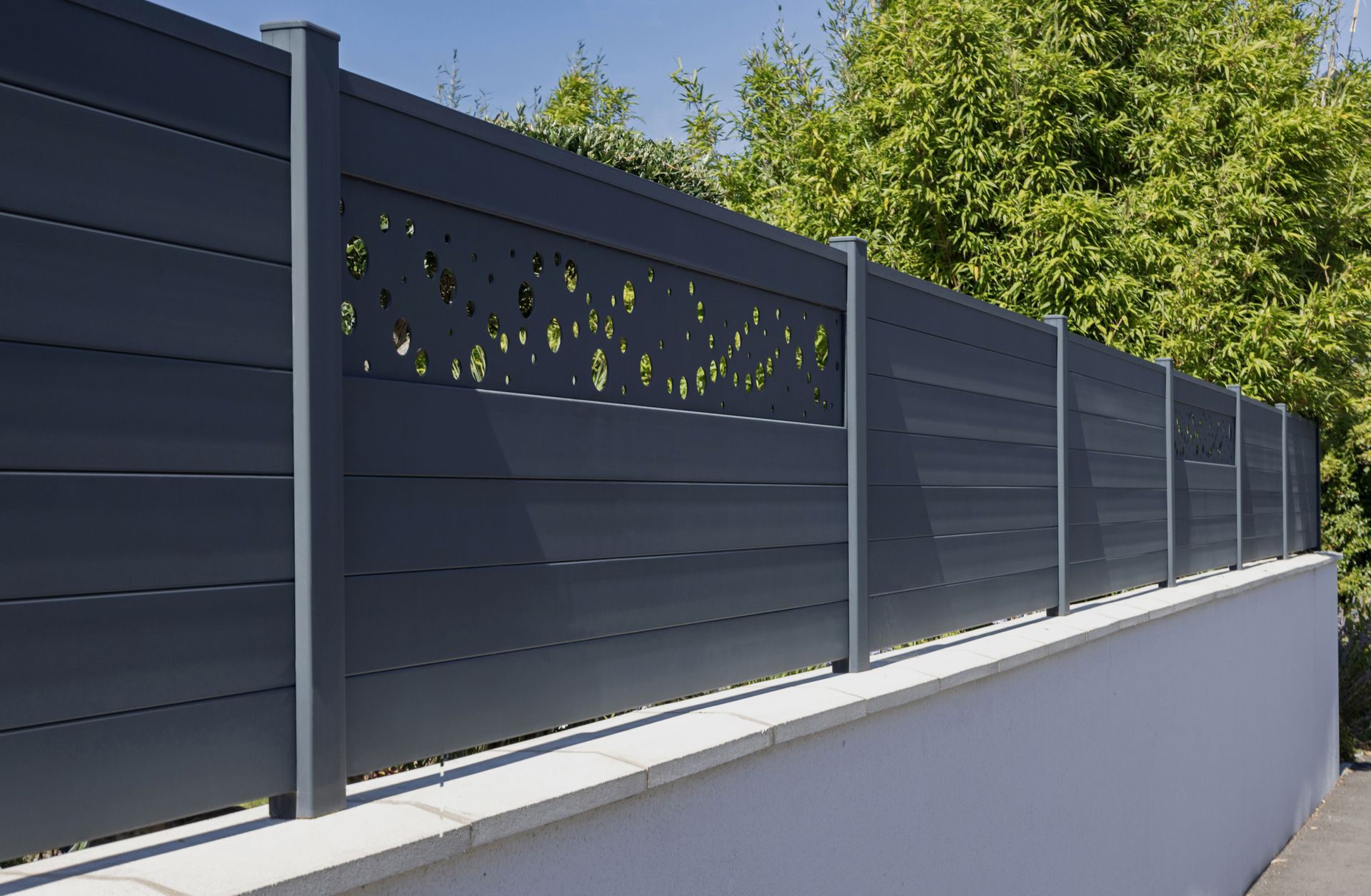 Clôture moderne gris foncé préservant l'intimité, ornée de découpes décoratives, le long d'un mur blanc bordé de verdure.