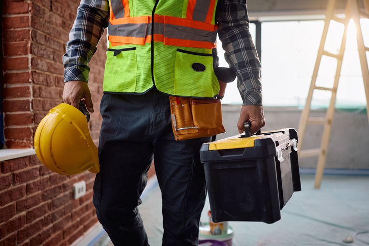 Ouvrier du bâtiment portant un gilet de sécurité, une caisse à outils et un casque jaune, sur un chantier.