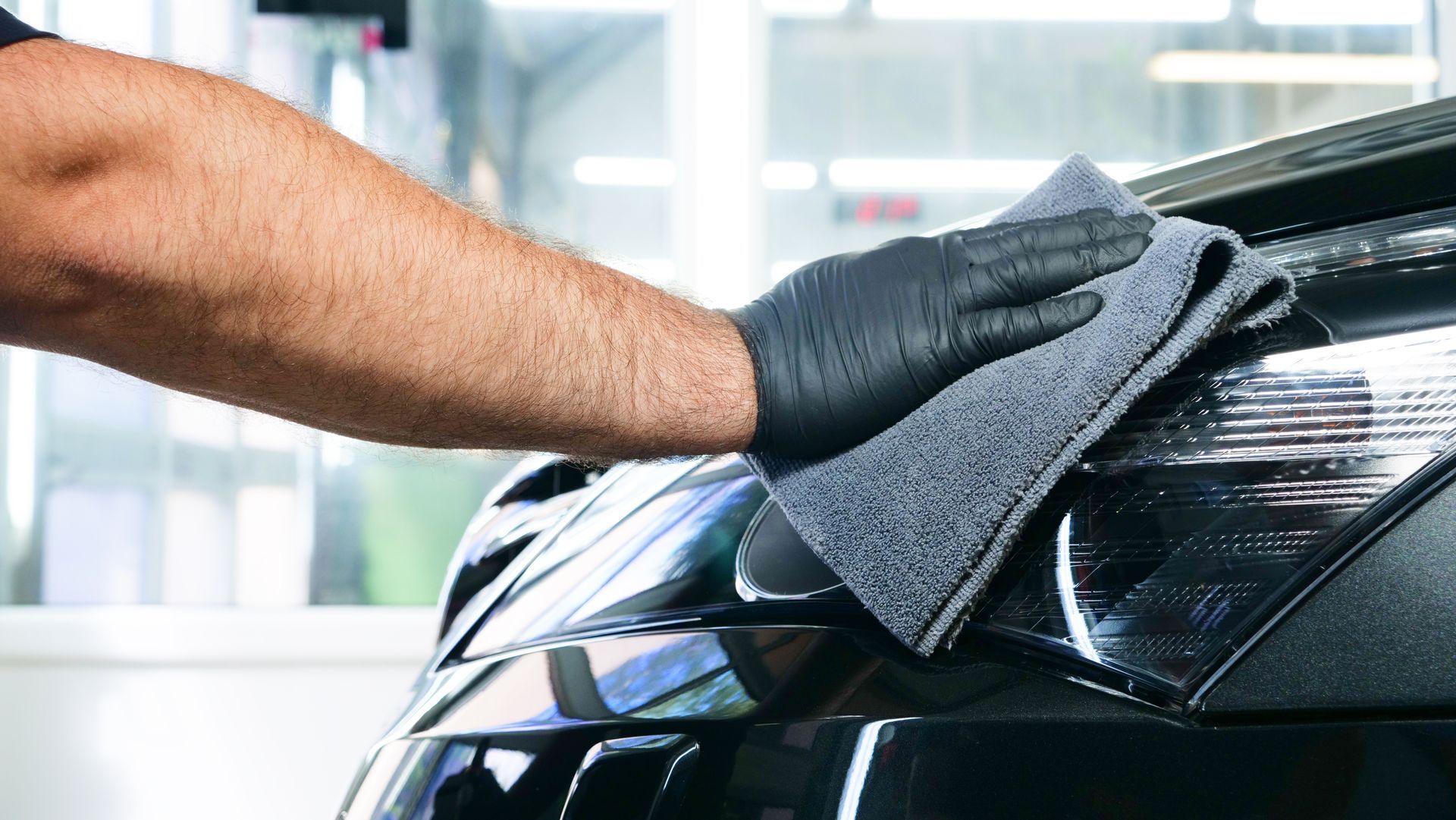 Main gantée de gris essuyant le capot noir d'une voiture avec un chiffon en microfibre.