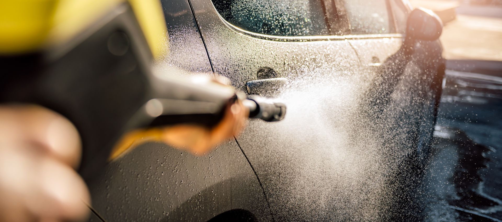 Une personne nettoie une voiture au nettoyeur à haute pression, projetant de l'eau et de la mousse sur une surface ensoleillée.