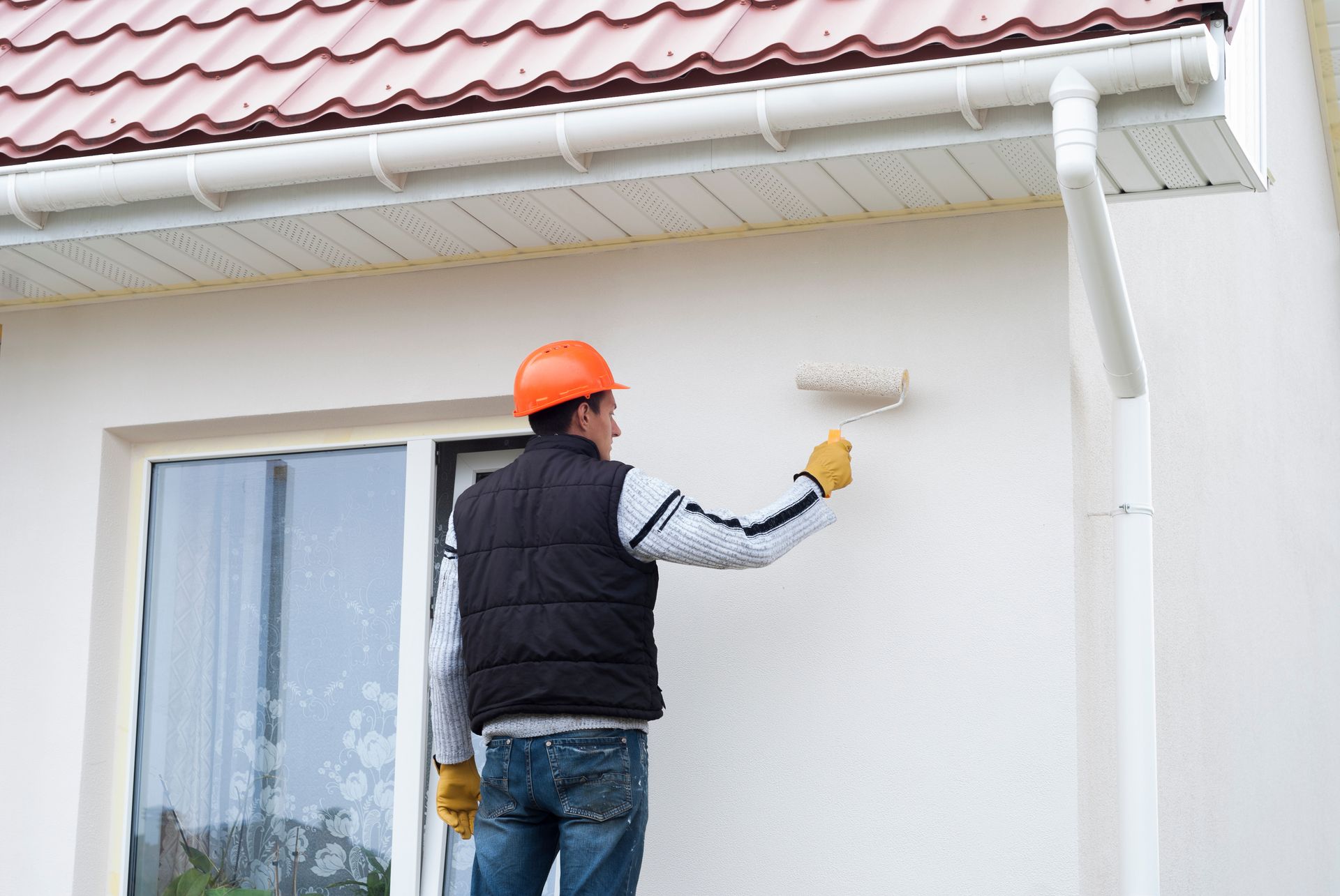 Un ouvrier portant un casque orange peint en blanc le mur extérieur d'une maison.