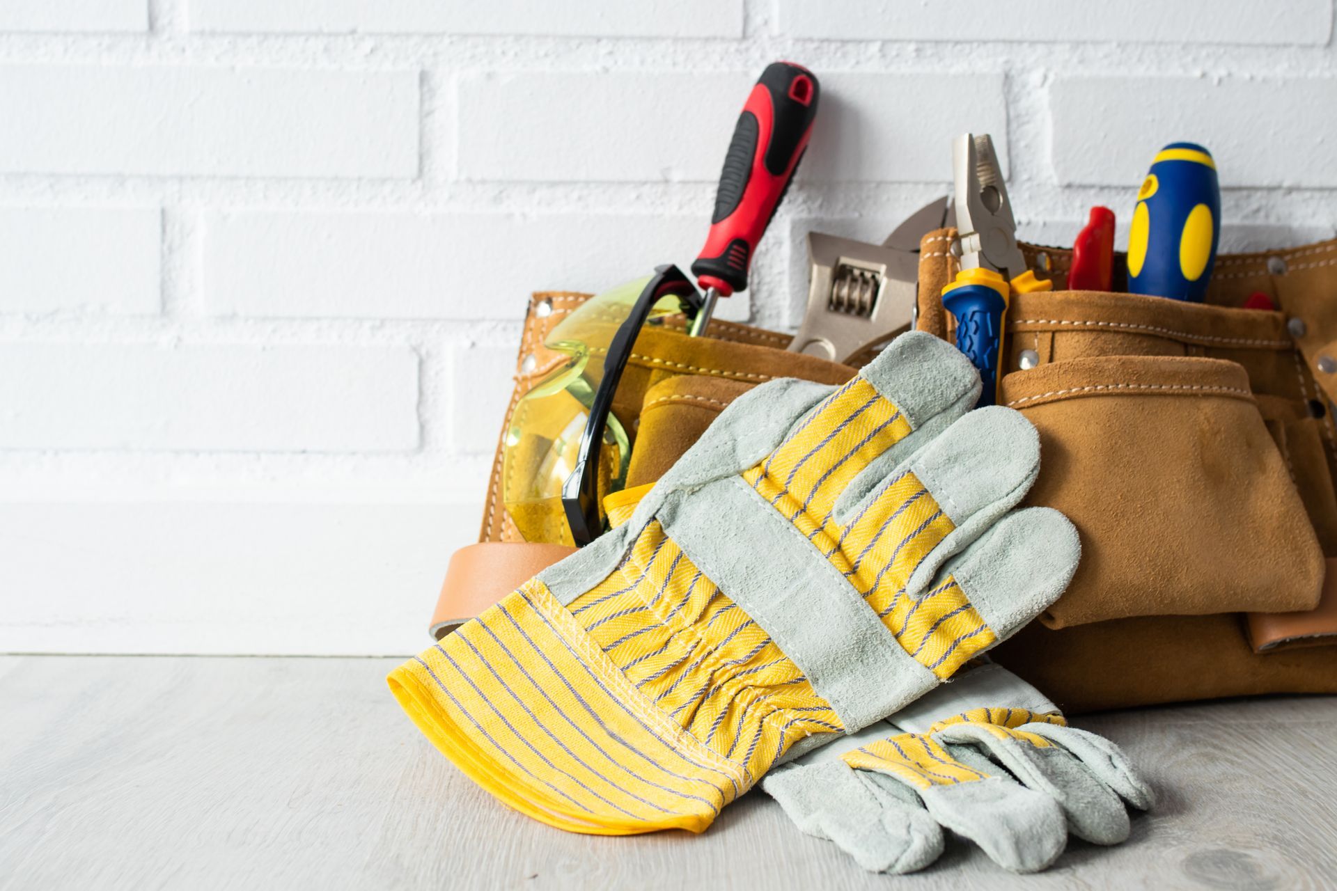 Gants de travail et outils à main dans une sacoche marron contre un mur de briques blanches.