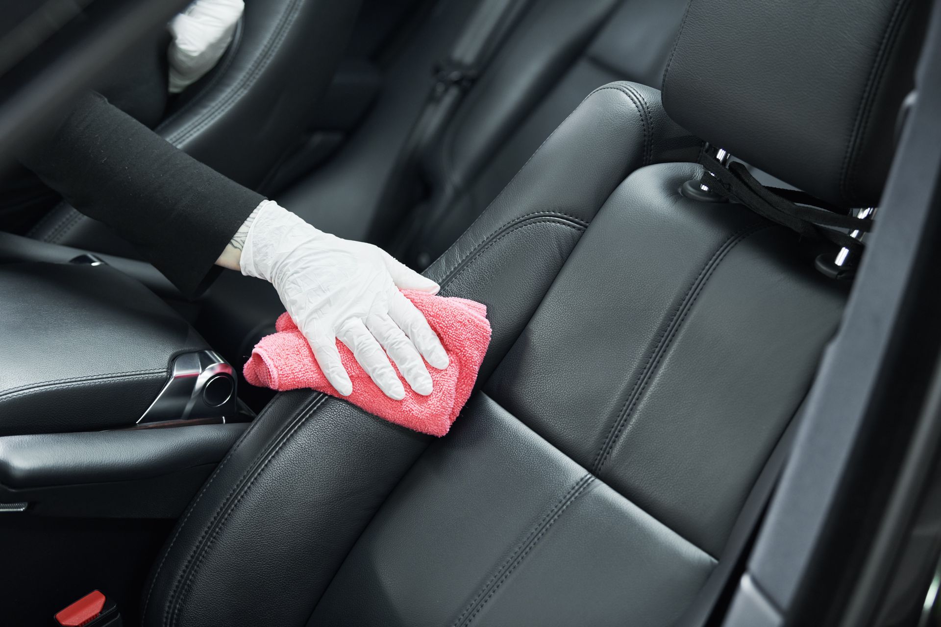 Main gantée essuyant un siège auto noir avec un chiffon rose à l'intérieur d'un véhicule.
