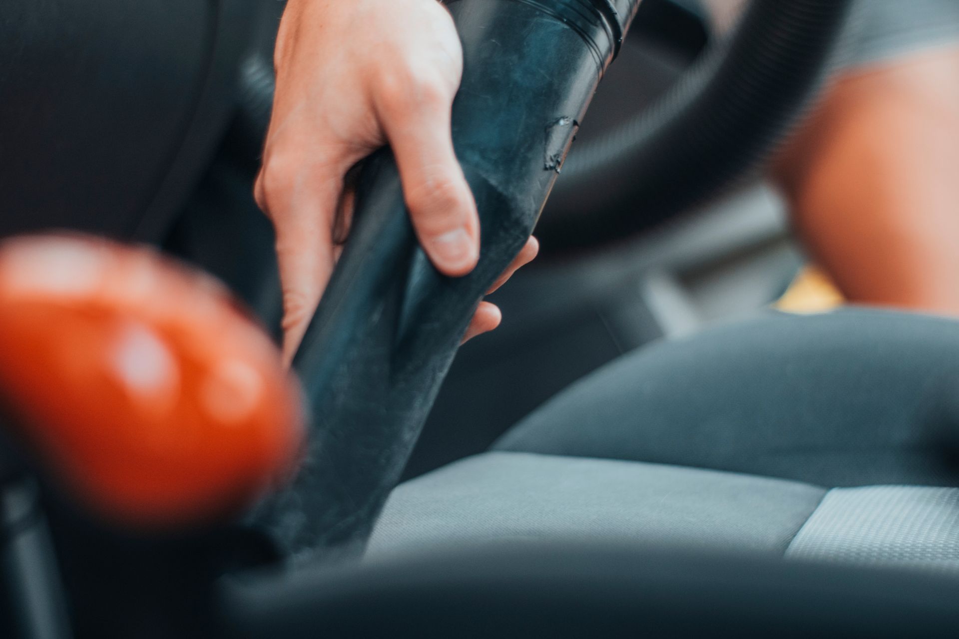 Aspirateur manuel nettoyant le revêtement d'un siège auto avec un objet rouge flou au premier plan.