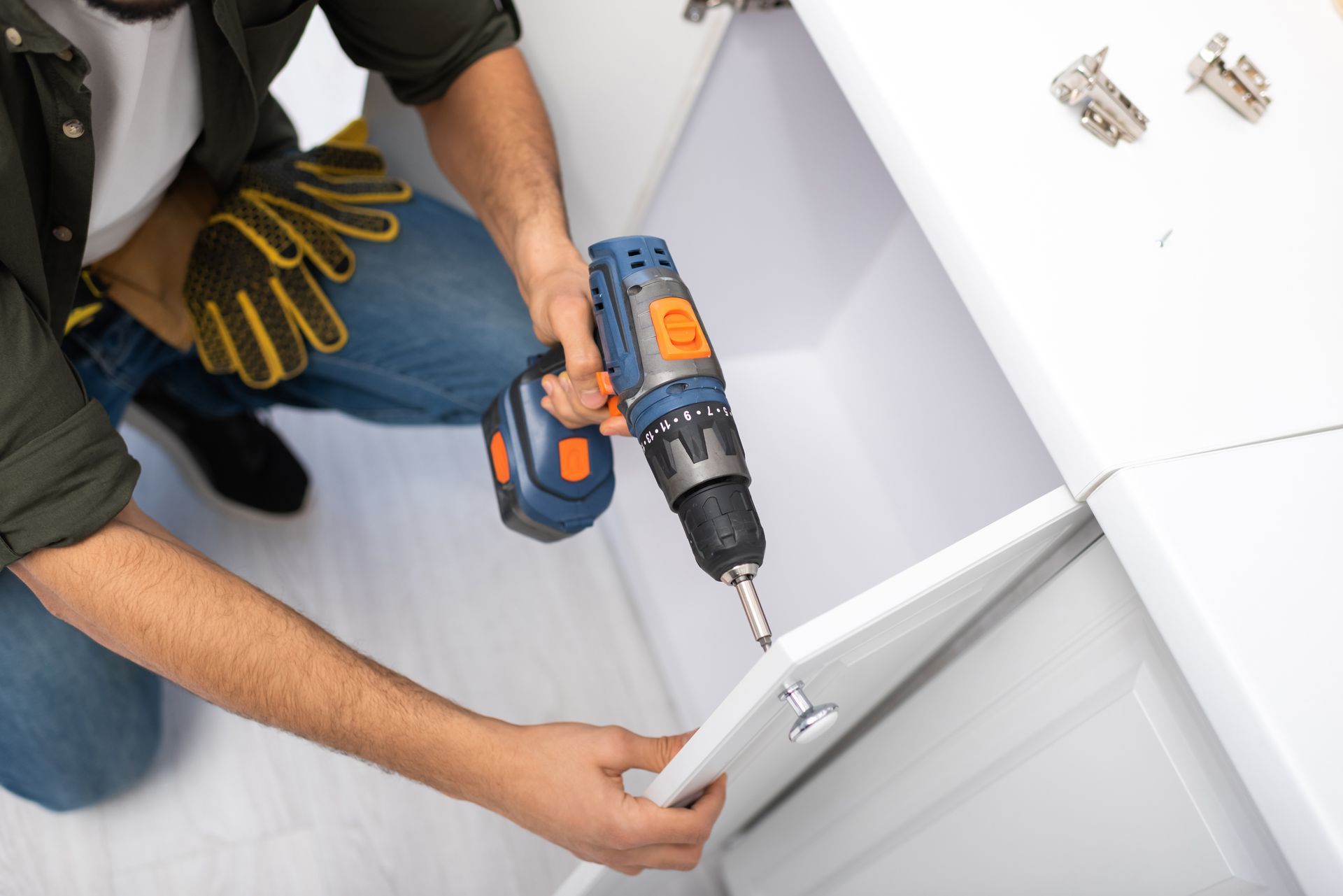 Une personne utilise une perceuse sans fil pour installer une porte d'armoire blanche dans un meuble de cuisine.