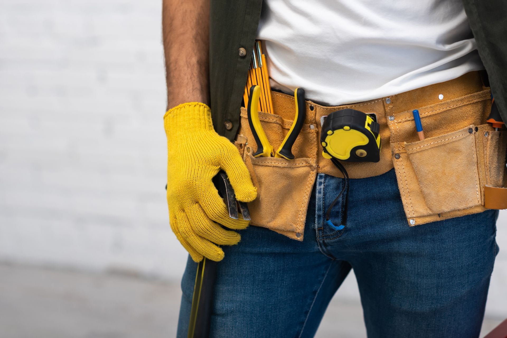 Ouvrier du bâtiment portant des gants jaunes et une ceinture à outils, tenant un outil contre un mur blanc.